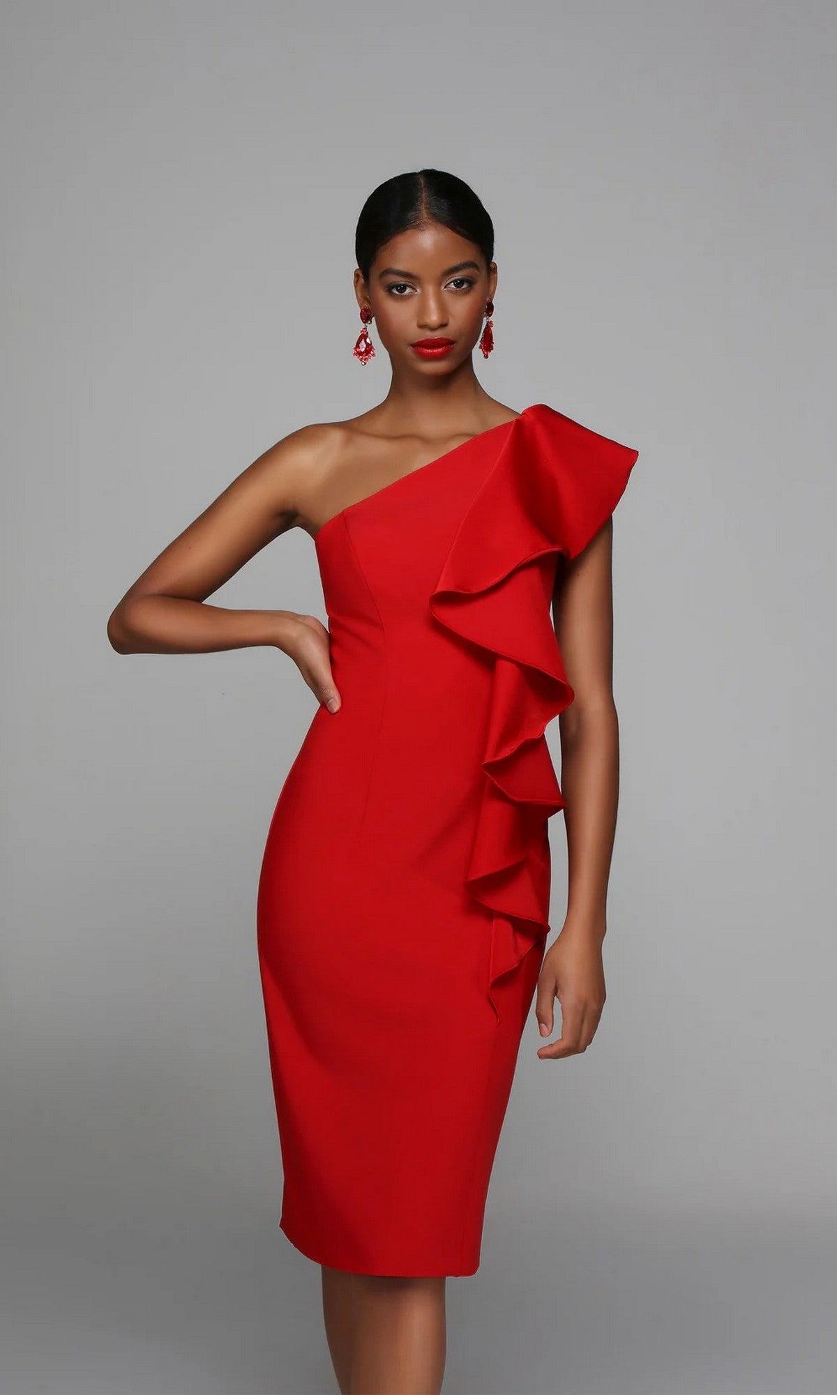 A woman models the Alyce 70008 Knee Length Semi-Formal Dress in red, featuring a fitted one-shoulder design and ruffle detail on the right side, standing before a plain gray background.